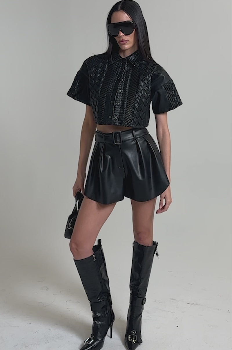 A video of a woman wearing the BOUNCE IT LIKE THAT FAUX LEATHER SHORT, a black crocodile-pattern crop jacket, knee-high boots, sunglasses, and holding a small handbag.