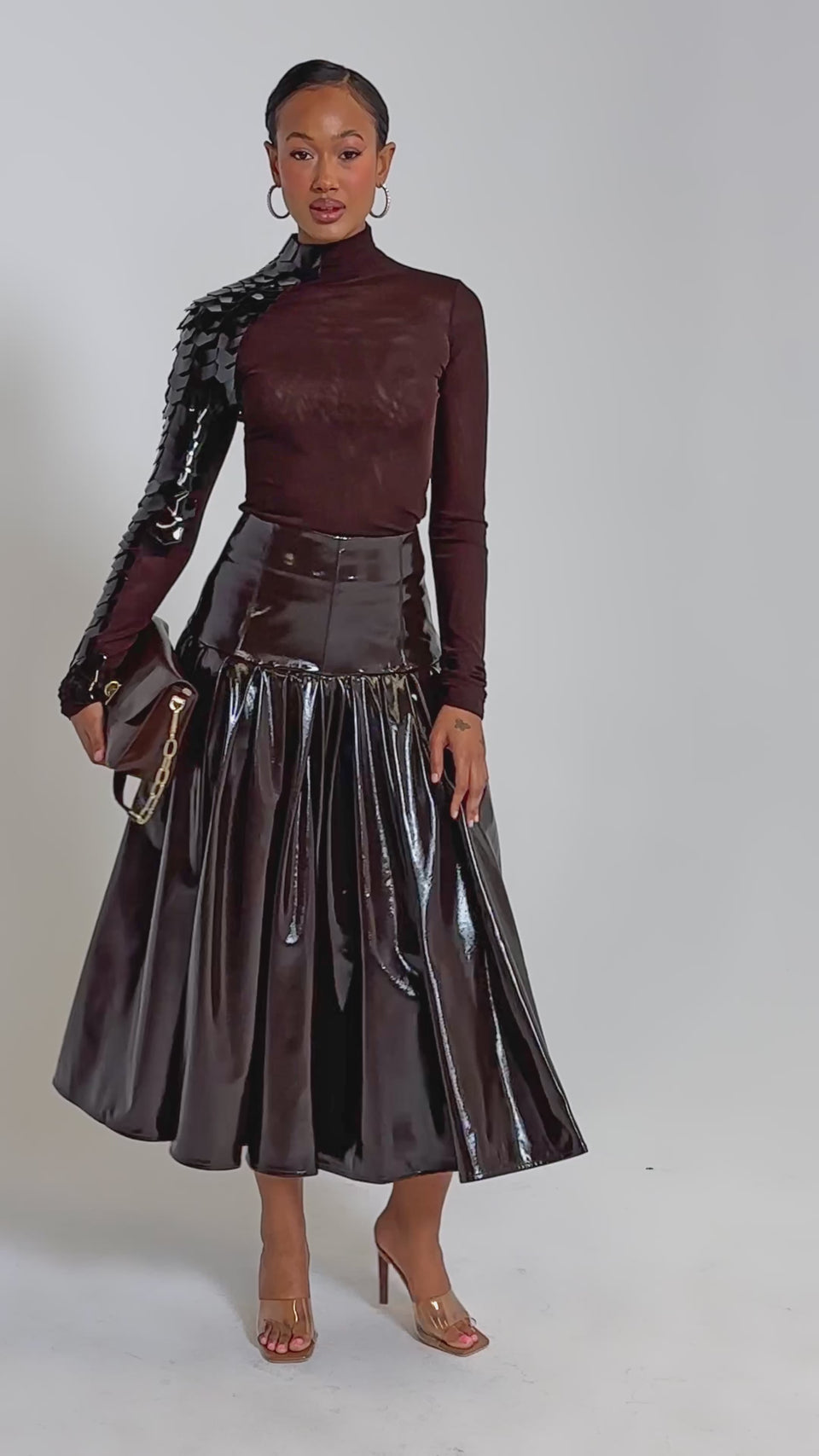 A video of a woman wearing a shiny brown midi skirt, the VELMA VINYL SINGLE SLEEVE SCALE TOP IN BROWN, clear high heels, and holding a brown clutch with a gold chain.