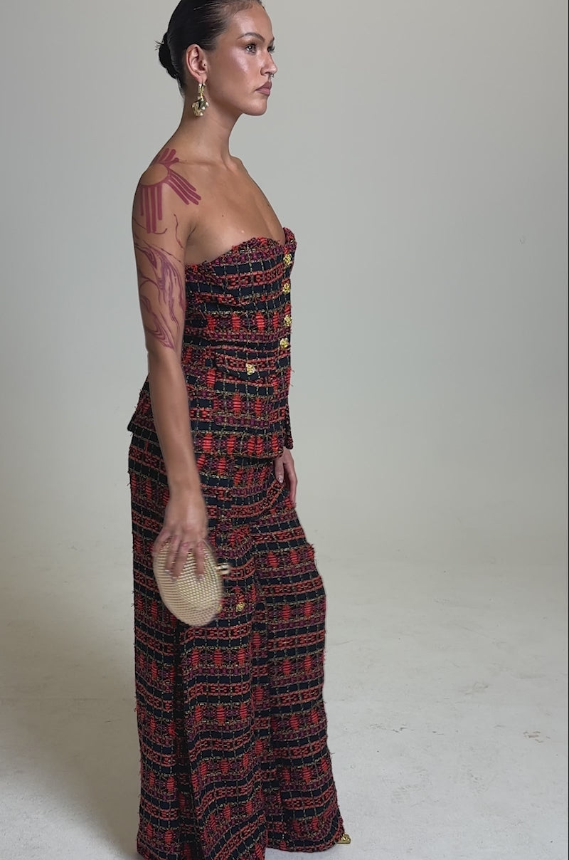 A video of a model wearing the ONLY THINK ABOUT YOU TROUSER IN TWEED, a wide-leg red and black patterned jumpsuit. She accessorizes with gold earrings, a beige clutch, and displays tattoos on her arms.