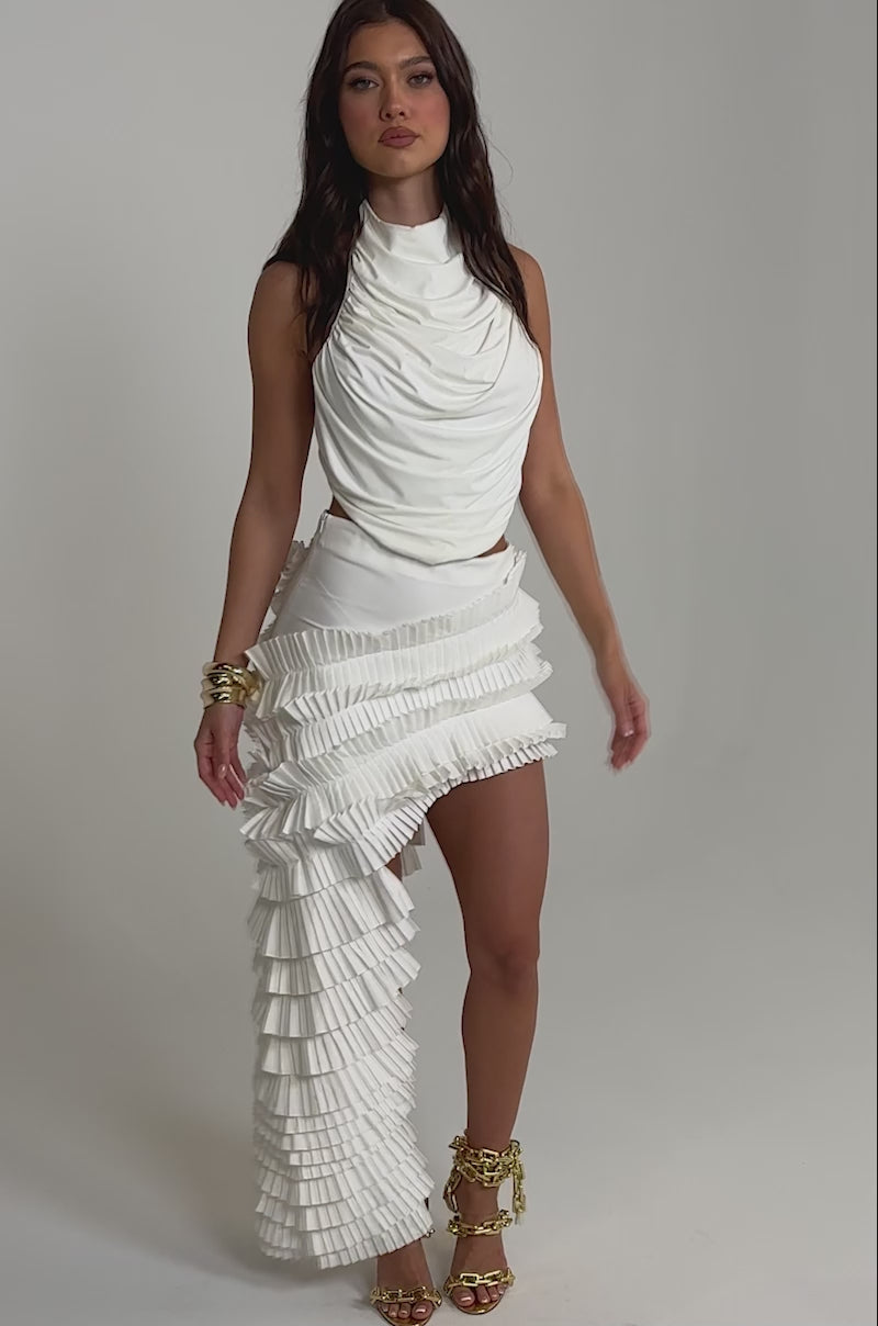 A video of a woman poses against a plain background wearing the DANCE THE NIGHT AWAY RUFFLE SKIRT, styled with a white sleeveless draped top, strappy gold heels, gold bracelets, and a small white bag.