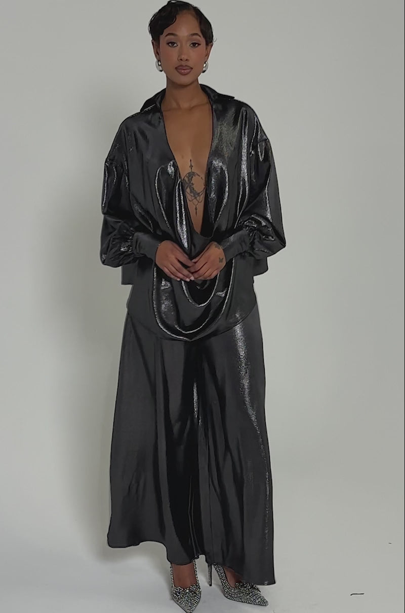 Video of woman modeling the MELINIA METALLIC WIDE LEG PANT, featuring glossy metallic fabric and a high-rise fit. She pairs it with a deep cowl neckline top, silver choker, and sparkling heels against a light gray background.