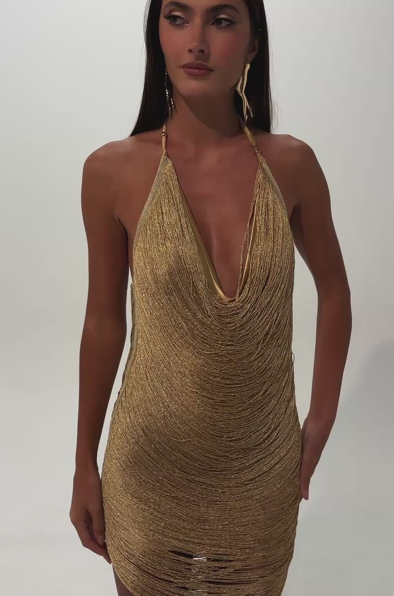 A video of a woman with long brown hair models the MATCHING ENERGY METALLIC FRINGE MINI DRESS IN GOLD—a shimmering gold halter mini with a draped neckline.