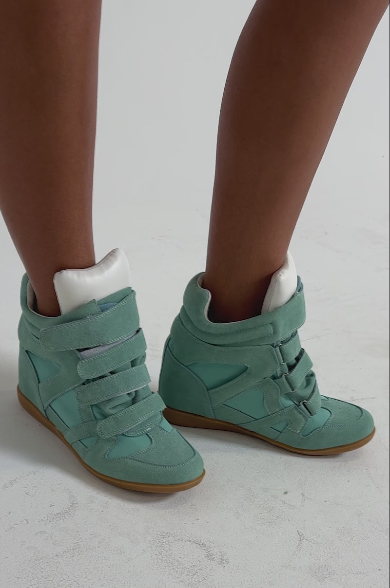 A video of a person wearing the AZALEA WANG BRYANT TEAL WEDGE SNEAKER with velcro straps and gum soles, standing with legs crossed against a plain, light background.