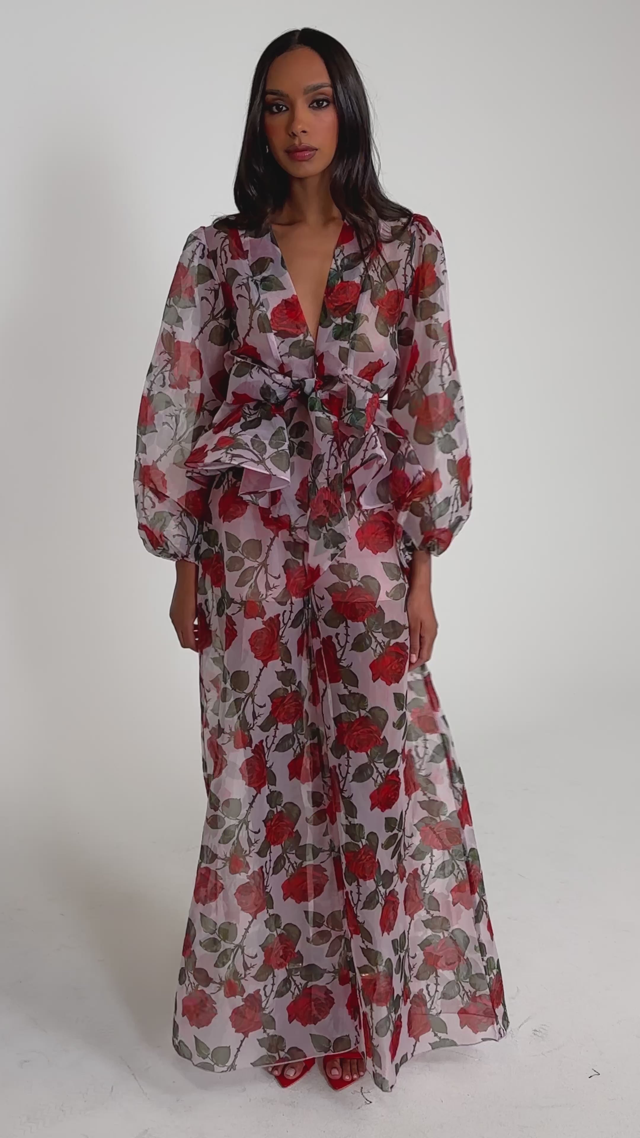 Video of woman modeling the PRETTY GIRL FLORAL TROUSER—a sheer, long-sleeved pink, red, and green rose print trouser with high-rise bow waistband and wide legs—paired with red heeled sandals against a plain light gray background.
