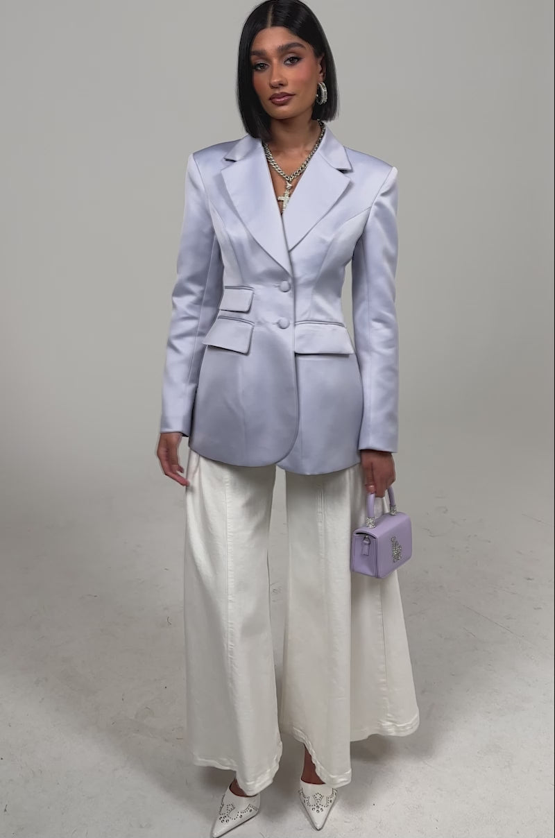 Video of a woman modeling the PERFECT FIT BLAZER IN LILAC, featuring two buttons and flap pockets, paired with white pants and accessories, standing against a plain light background.