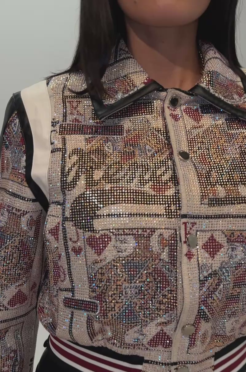 A video of a woman wearing oversized sunglasses and the RAISE YOUR BET RHINESTONE BOMBER—a jacket featuring a playing card graphic with red, black, and white trim.