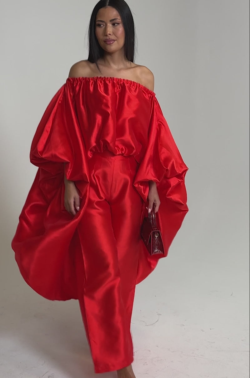 A video of a woman wearing vibrant high-rise wide-leg BY MY SIDE TROUSER pants with a red satin off-the-shoulder top, dramatic sleeves, a small red purse, and red high-heeled sandals.