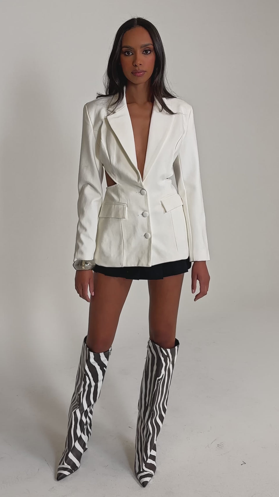 Video of woman modeling the PARTY IN THE BACK CUTOUT BLAZER IN WHITE over a black mini skirt, paired with layered jewelry, posing against a light background—perfect for modern office-wear with an edgy vibe.