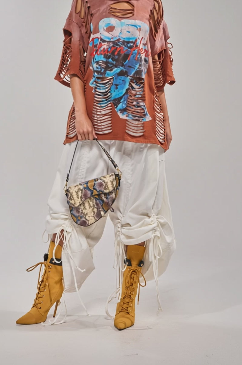A video of a person in the DRAMA RUCHED JOGGER IN WHITE, paired with mustard yellow lace-up boots, a cropped rust top, and a snakeskin-print handbag against a plain background.