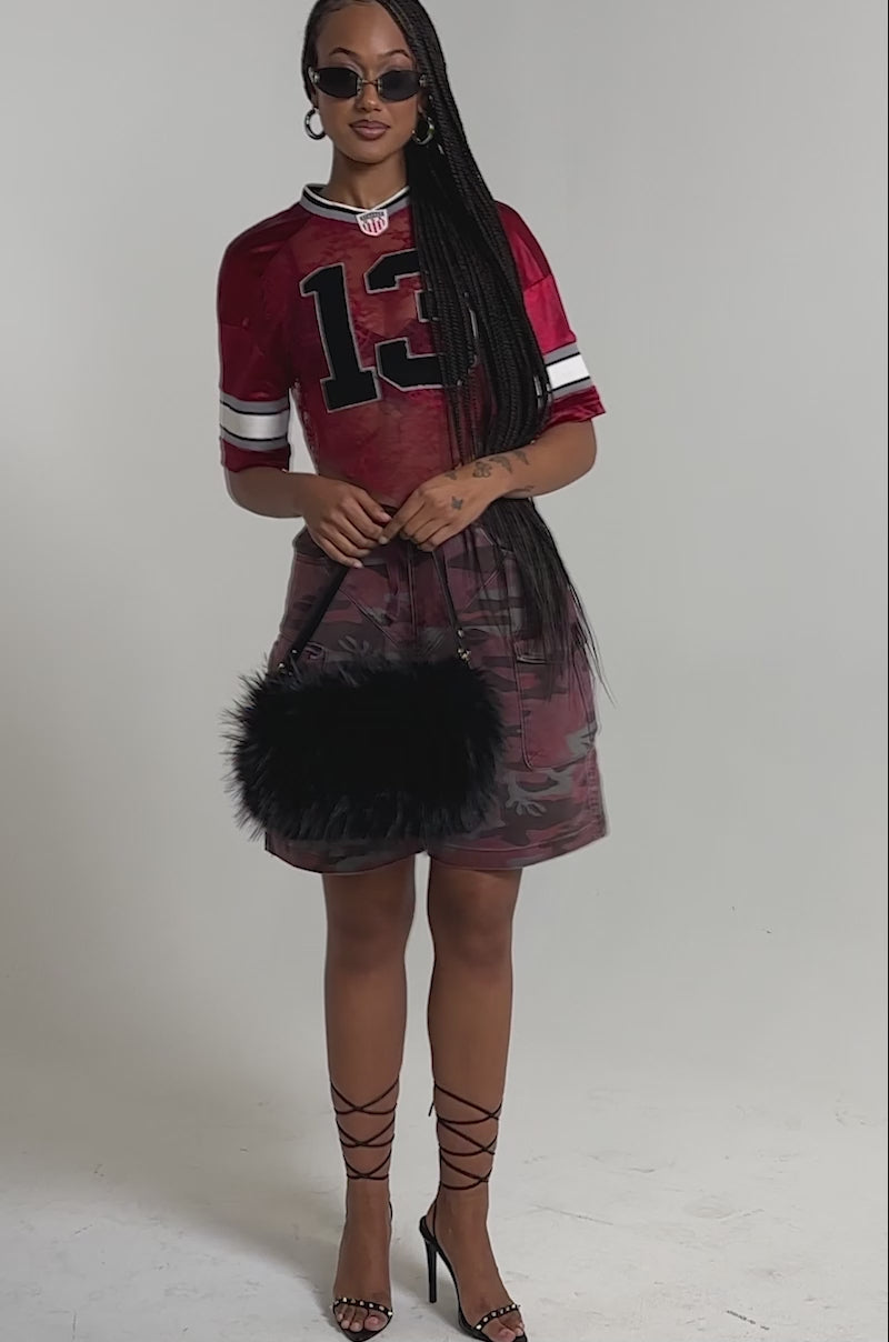 A video of a woman in the LUCKY LACE JERSEY TOP IN BURGUNDY with camo shorts, strappy heels, sunglasses, hoop earrings, and a black furry handbag.