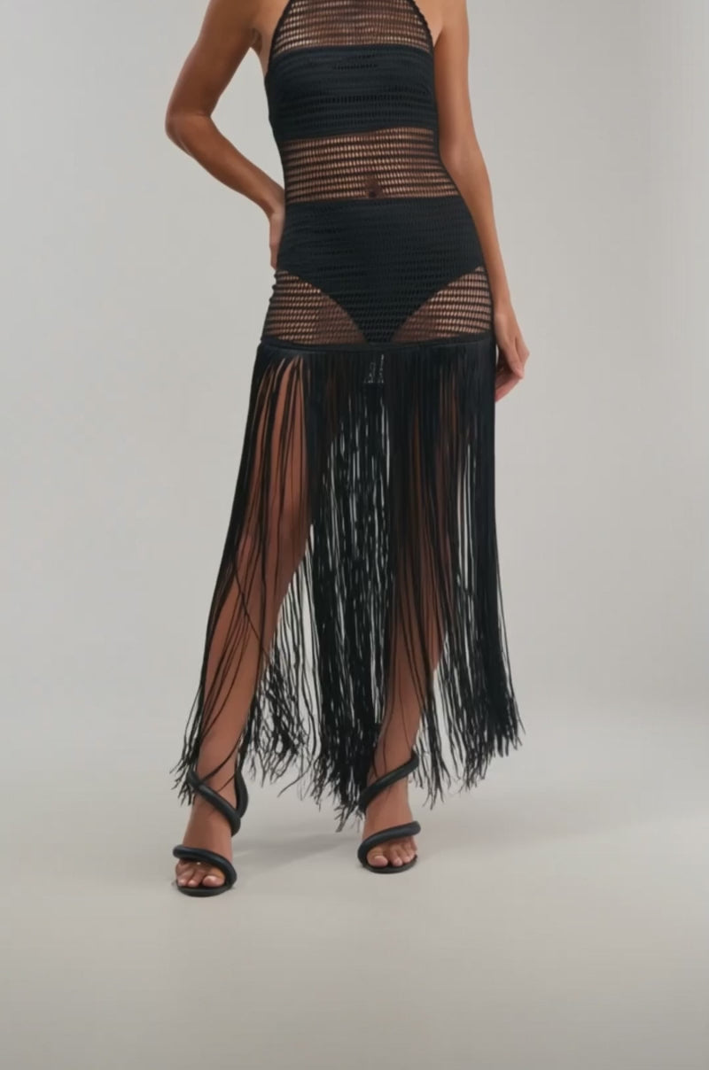 A video of a woman in the OCEAN BREEZE HIGH NECK FRINGE MAXI DRESS, a sleeveless black crochet beach dress with see-through panels and long fringe, paired with black sandals and posing against a plain background.
