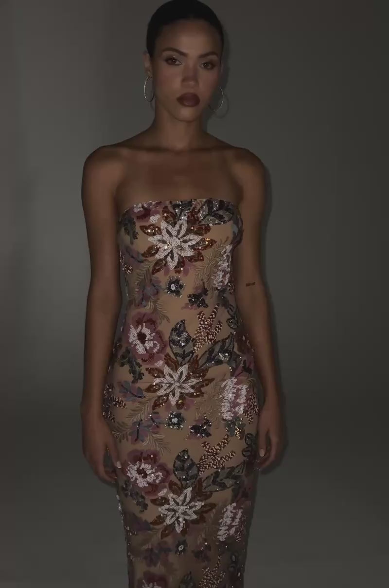 Video of woman modeling the SHOW ME LOVE EMBELLISHED MIDI DRESS, a strapless sequin midi with a nude base and vibrant floral embroidery, styled with transparent heels. She stands against a plain gray background, highlighting this elegant wardrobe piece.