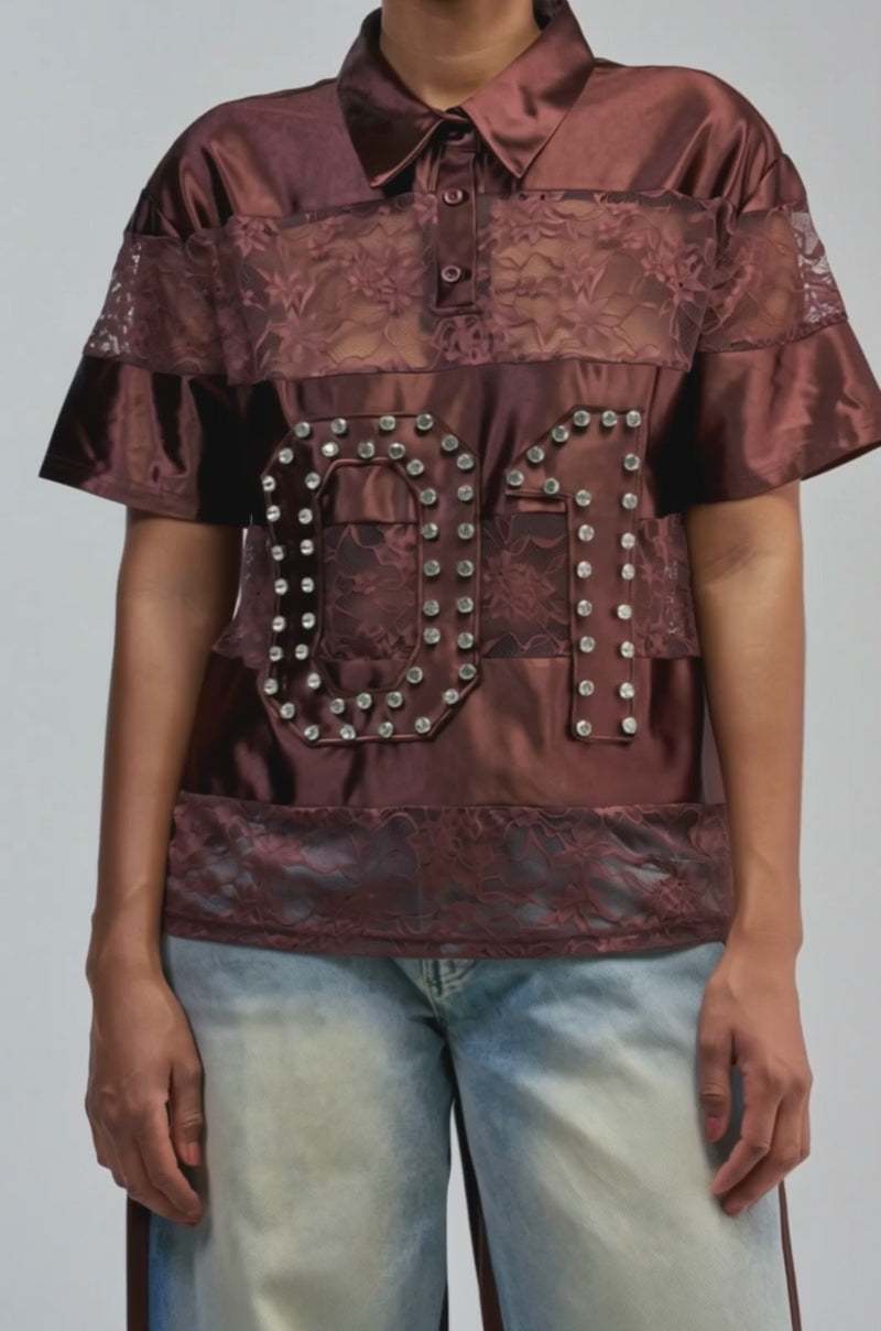Video of a woman modeling the NEW AUTHORITY LACE SHORT SLEEVE POLO—a lustrous burgundy shirt featuring sheer floral lace accents and a “01” motif outlined in metal studs.