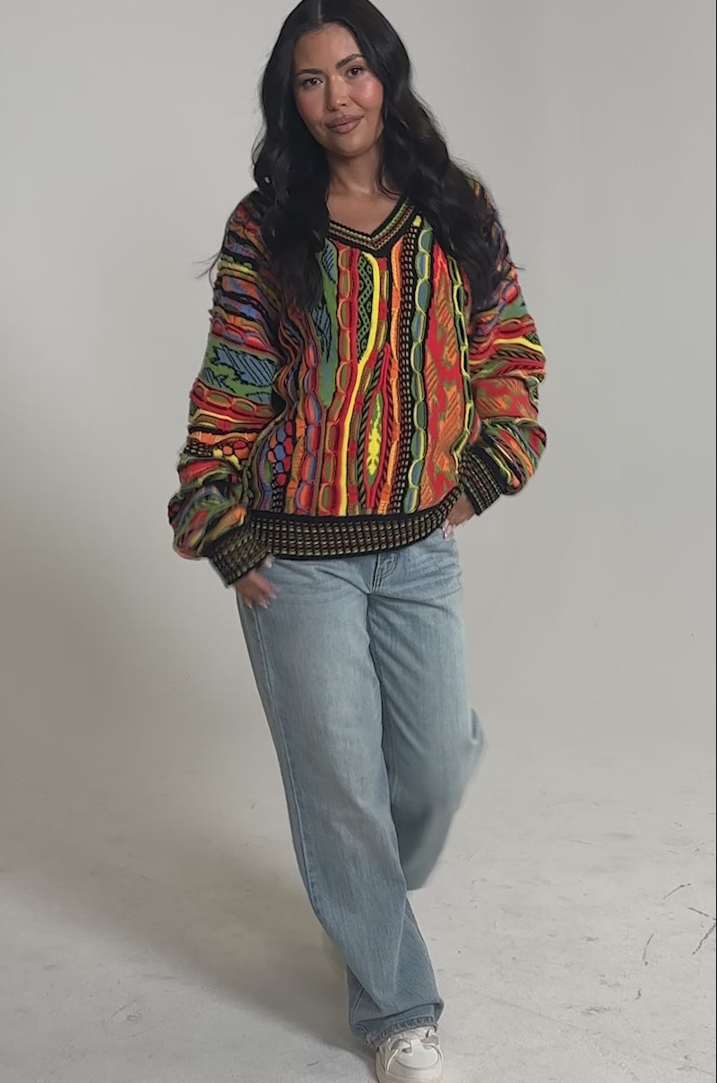 Video of woman modeling the COSMIC WEAVE V-NECK SWEATER, featuring bold multicolor patterns and loose sleeves, paired with light blue jeans against a plain light background.