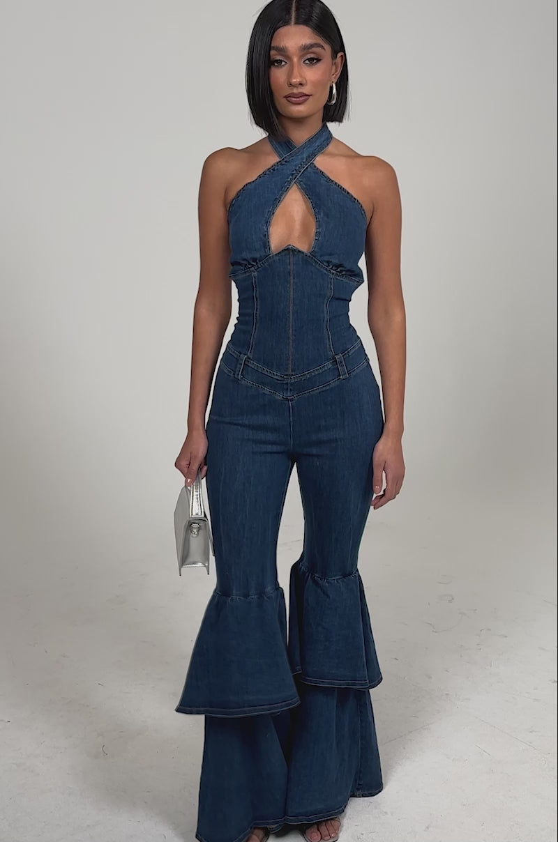 Video of woman modeling the RODEO REBEL DENIM JUMPSUIT with a keyhole cutout, fitted waist, and flared ruffle legs. She accessorizes with a small silver handbag and strappy heels for a chic, retro-inspired style.