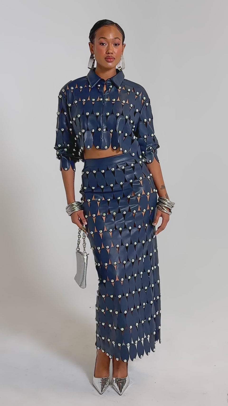 A video of a woman in THE SNEAK PEEK STUDDED PU CROP CARDIGAN IN NAVY with oval cutouts, styled with a matching long skirt, silver purse, silver heels, and statement jewelry against a plain backdrop.