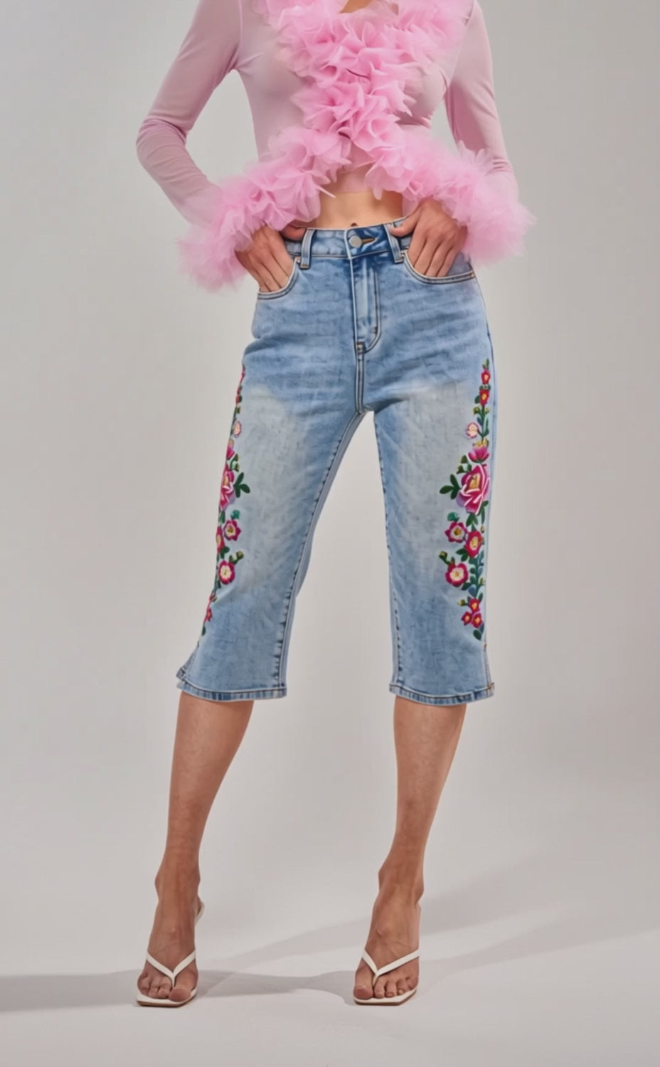 Video of woman modeling the GIVE ME FLOWERS FLORAL CAPRI—floral embroidered, cropped denim capris—paired with clear strap high-heeled sandals and a sheer pink ruffled top, set against a plain light background.