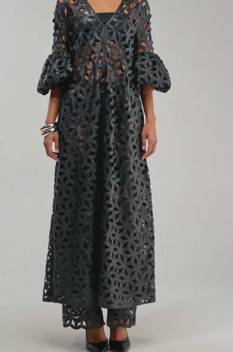 A video of a woman wearing the LEAH DUSTER IN BLACK—a longline statement piece—over a long, sheer black dress with geometric cutouts and voluminous three-quarter sleeves.