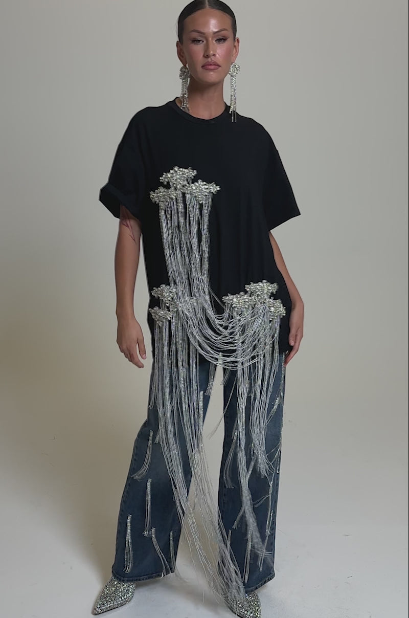 A video of a person wearing the SPARKLING OVERSIZED TEE—a black luxury cotton shirt with long rhinestone fringe and floral embellishments—paired with blue flared jeans and silver sparkling shoes, standing out against a neutral background.