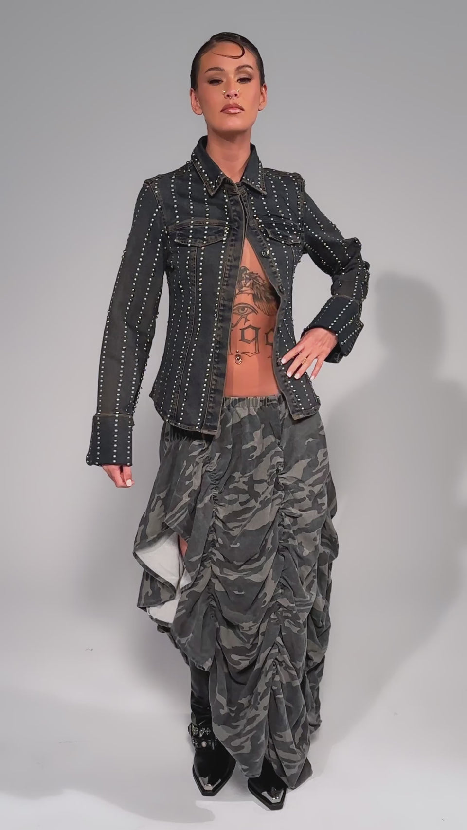 Video of woman modeling the LETS CHANGE IT UP RUCHED MIDI SKIRT IN CAMO, and tall black boots, posing against a plain light gray background.