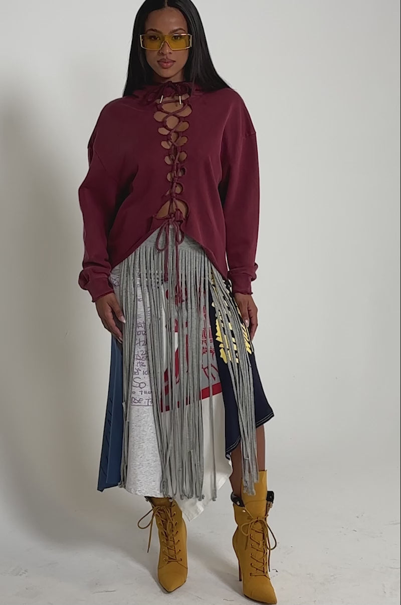 A video of a woman wearing in a burgundy lace-up sweatshirt, the STRESSED AND DISTRESSED MIDI SKIRT with fringe and graphic prints, yellow heeled boots, and yellow sunglasses.