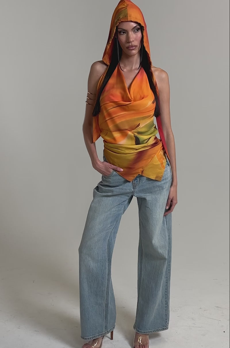 A video of a woman in the COASTAL HOODED HALTER TOP with orange and yellow draped fabric, styled with light blue wide-leg jeans and beige heeled sandals against a plain light background.