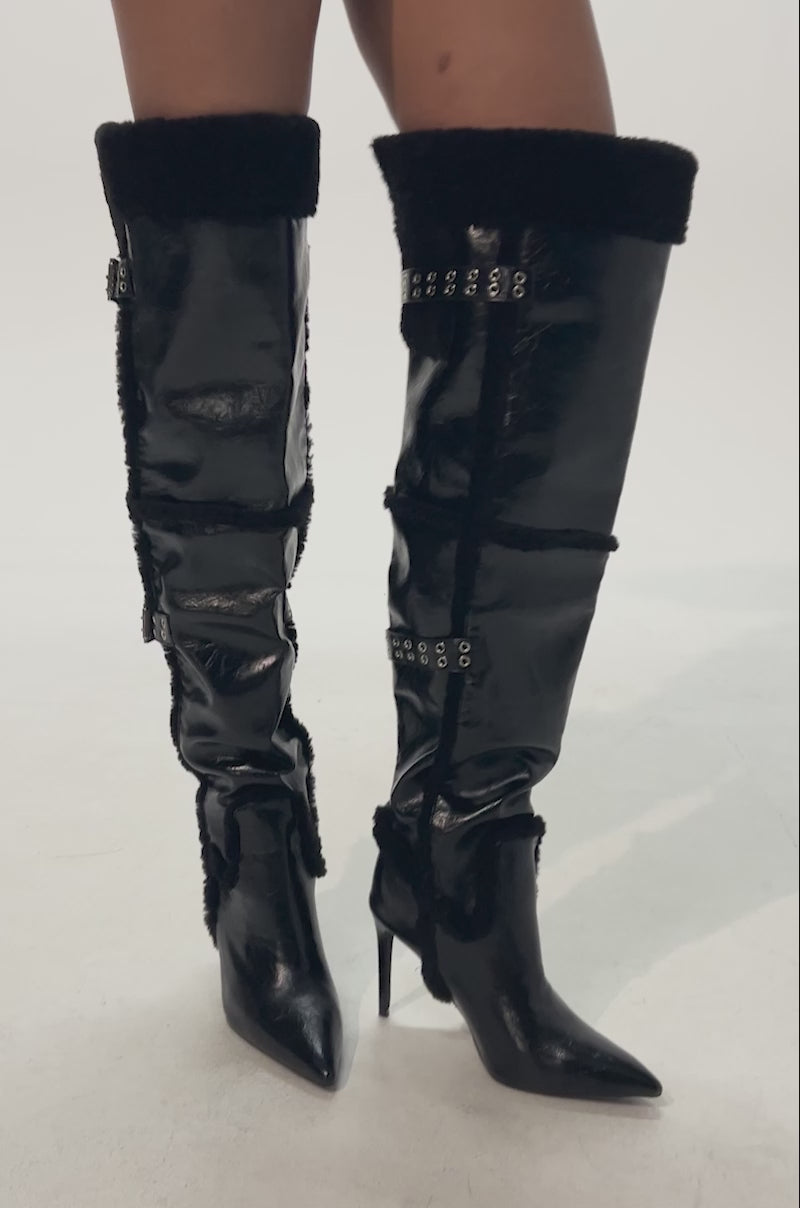 Video of woman modeling the AZALEA WANG XO SHERPA LINED PU STILETTO BOOTS—black knee-high faux leather with a shiny finish, shearling trim, decorative buckles, and cozy sherpa lining.