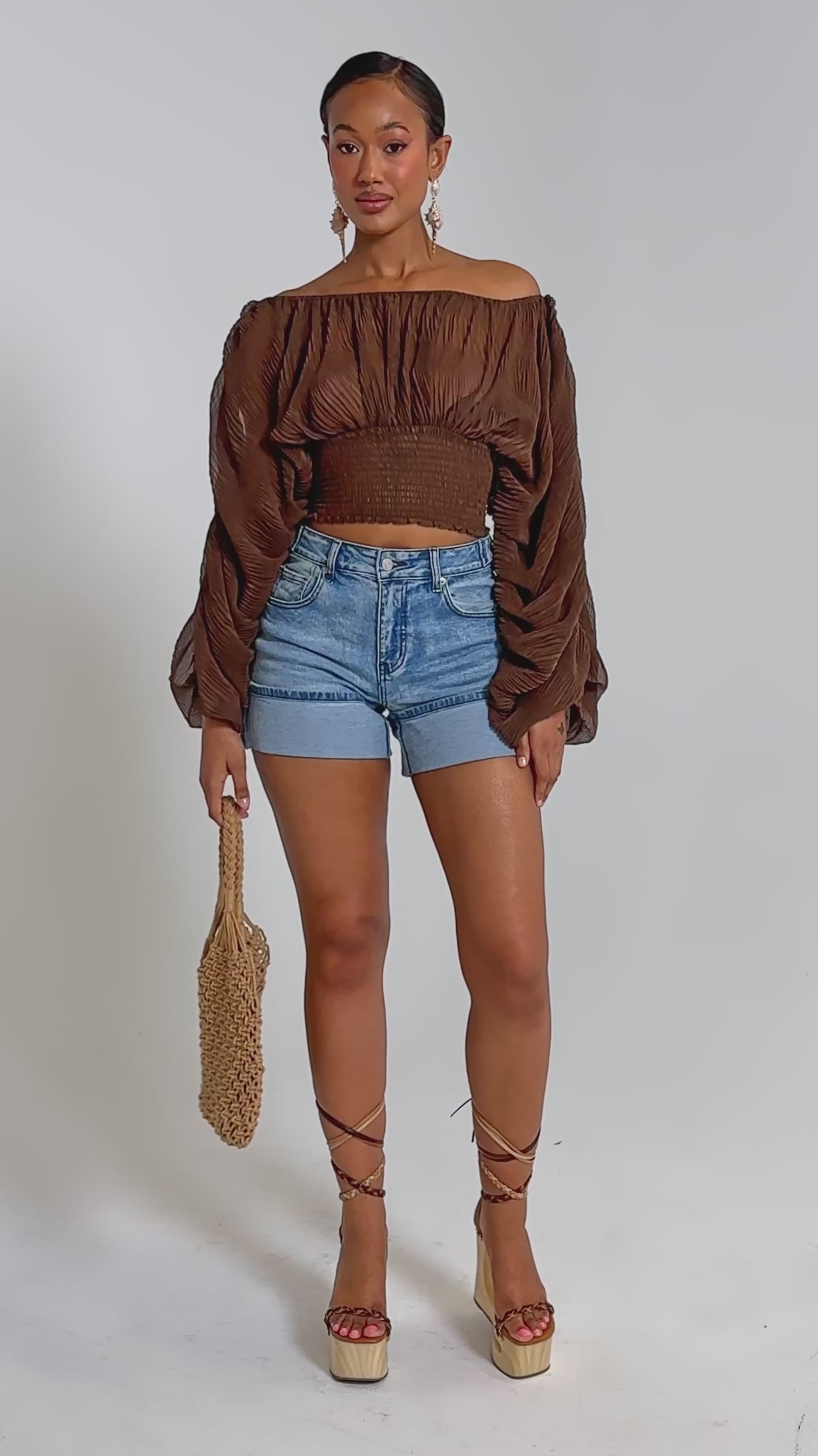A video of a woman wearing the DIVINE RIGHT LONG SLEEVE BLOUSE IN BROWN, paired with blue denim shorts, platform lace-up sandals, a woven straw bag, and hoop earrings.