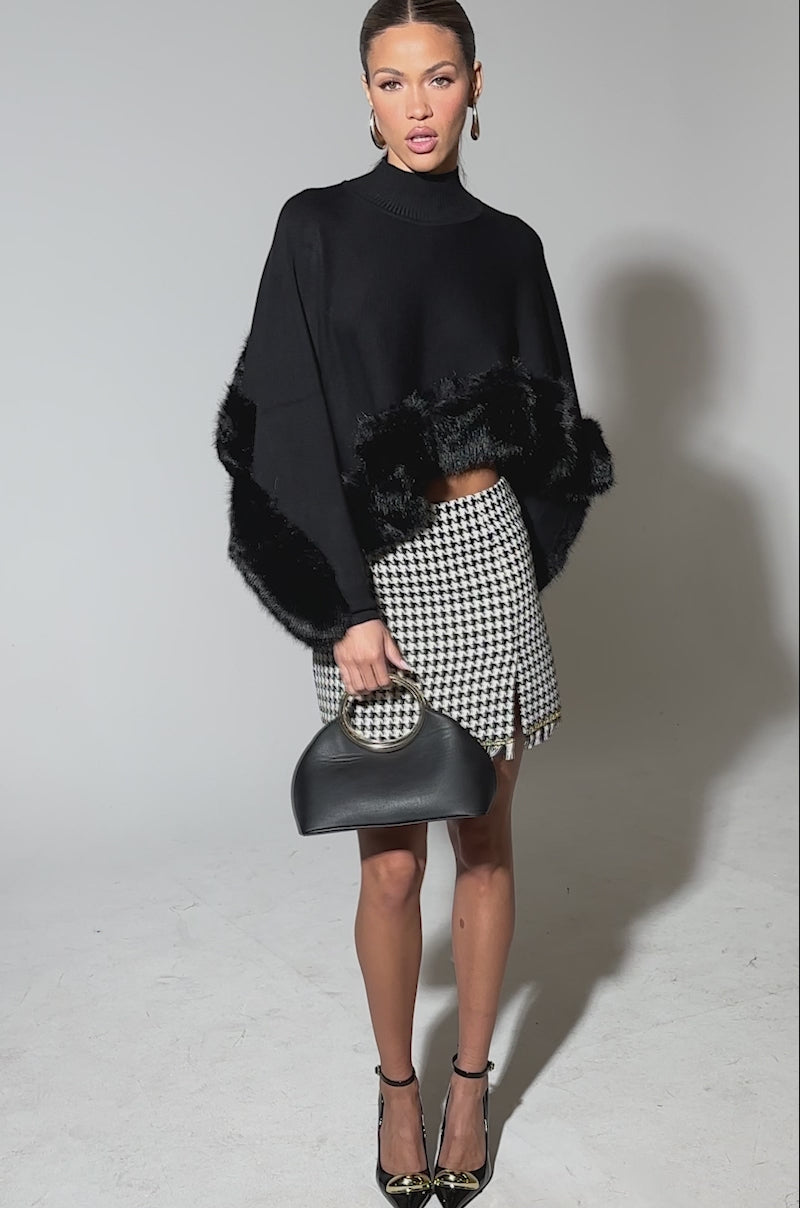 Video of woman modeling the QUIET LUX FUR TRIM SWEATER IN BLACK with a black-and-white houndstooth mini skirt with a slit, black heels featuring gold tips, and carries a black handbag with a gold handle.