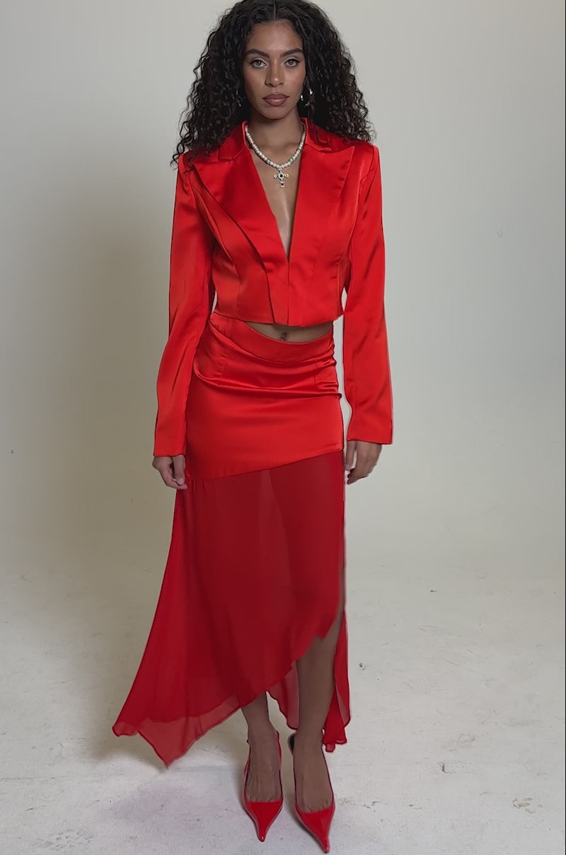 Video of woman modeling the RED HOT ASYMMETRICAL MIDI SKIRT with a cropped blazer, pointed heels, and layered necklaces, posing confidently against a plain light background.