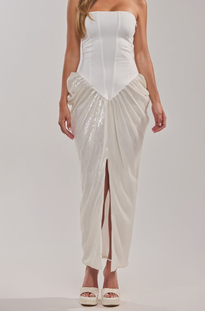 Video of woman modeling the GLOW GETTER SEQUIN CORSET MAXI DRESS with a structured, strapless bodice and a draped skirt with a front slit, she pairs platform heels and a necklace. Her long wavy brown hair completes the chic look against a plain background.