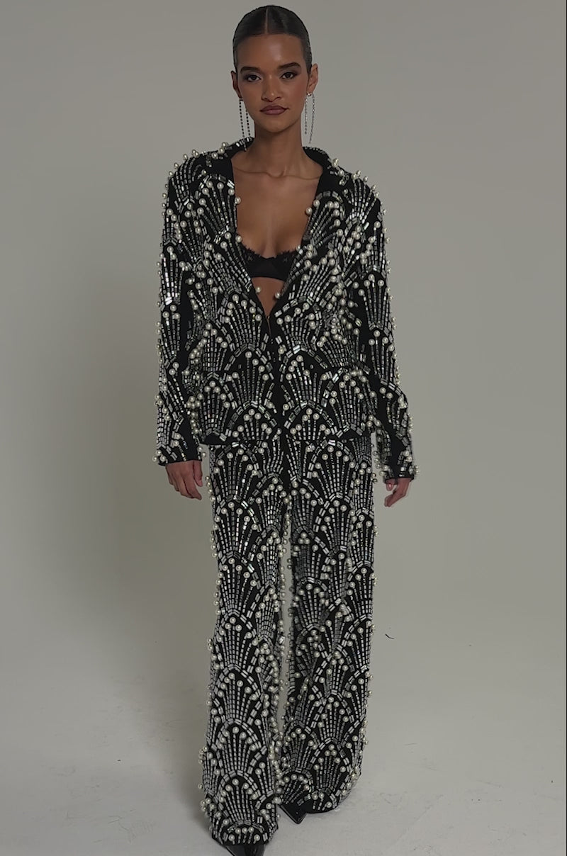 Video of woman modeling the TIMES SQUARE EMBELLISHED PANT, paired with a black suit featuring pearl and bead patterns over a black bralette, creating an elegant silhouette against a plain, well-lit background.