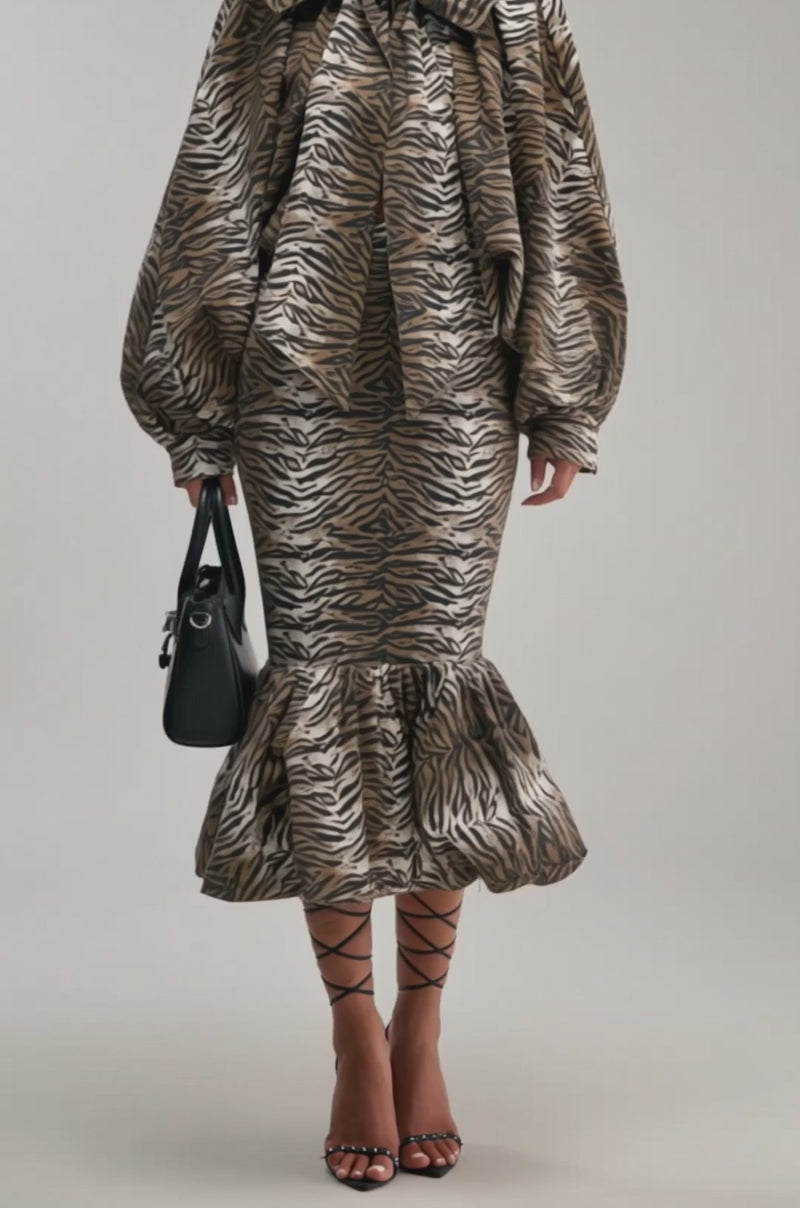 A video of a woman in the INTO THE WILD ZEBRA BLOUSE, paired with a ruffled skirt and strappy black heels, holding a black handbag against a light plain background.
