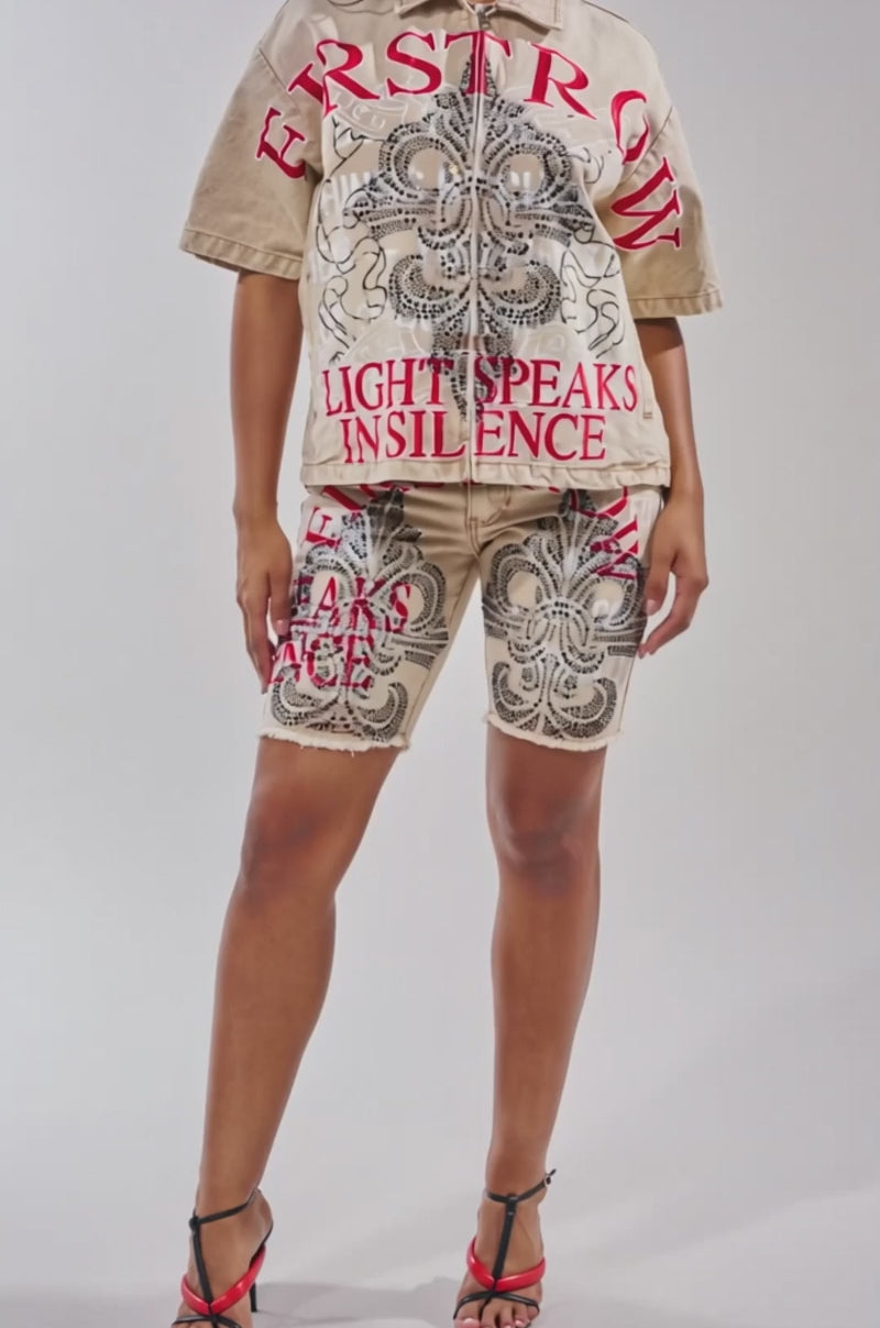 A video of a woman wearing the LIGHT SPEAKS IN SILENCE CANVAS SHIRT, a tan top with lace-like patterns, subtle rhinestones, and bold red text: "FIRST ROW" and "LIGHT SPEAKS IN SILENCE." She pairs it with matching shorts and strappy black and red heels.