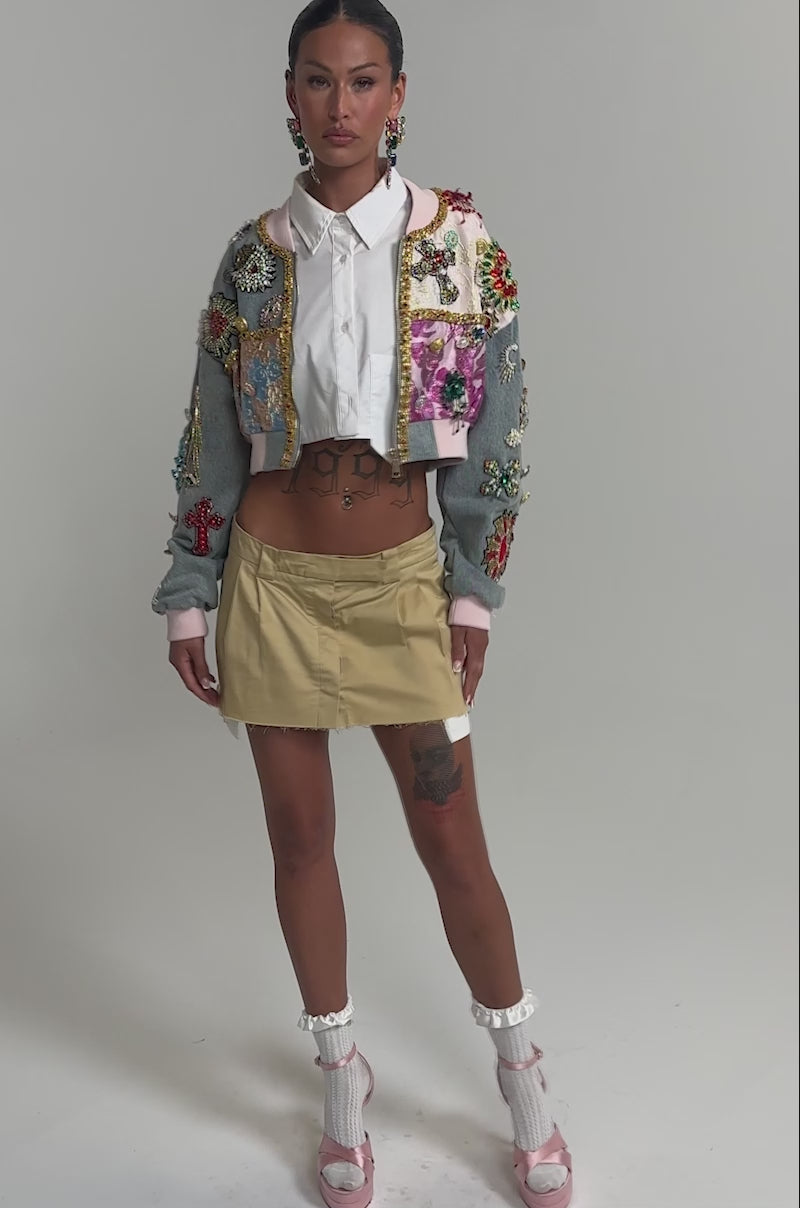 Video of woman modeling the IF YOU'RE DOWN EMBELLISHED DENIM BOMBER with colorful details, styled with a pleated tan mini skirt and statement earrings, against a plain background.