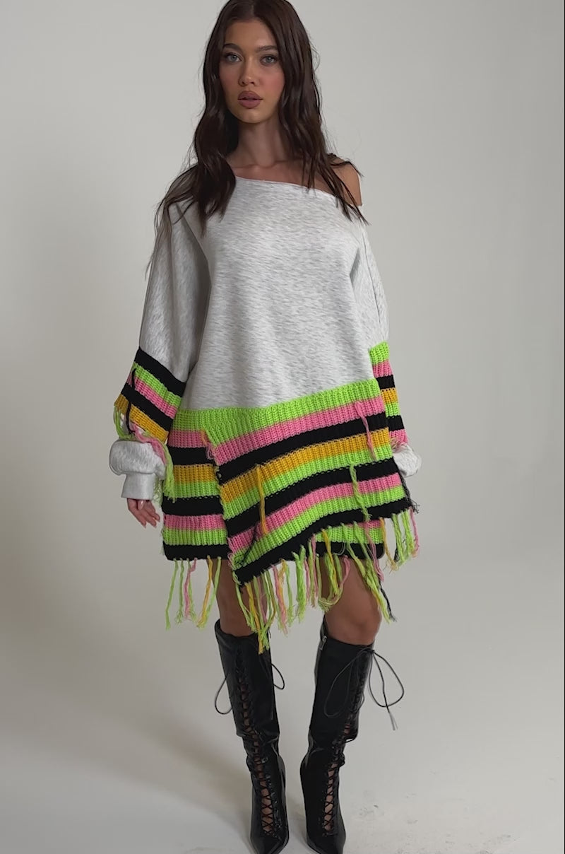 A video of a woman wearing the MOST LOVED SWEATSHIRT MINI DRESS featuring neon green, pink, and black stripes with crochet fringe, paired with knee-high black lace-up boots.