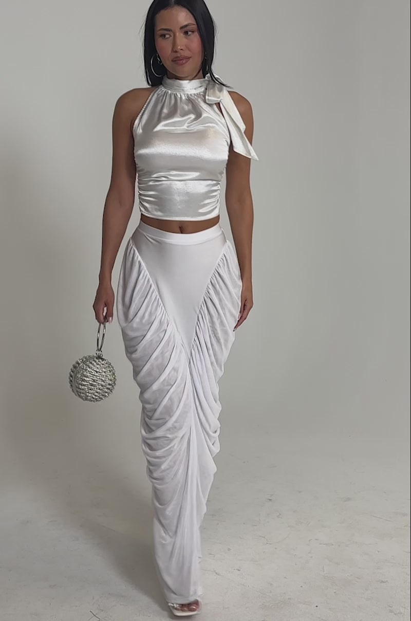 Video of woman modeling the DREAMY RUCHED SKIRT IN WHITE with a shiny silver sleeveless top and tied neck, holding a round beaded purse while smiling on a plain, light background.