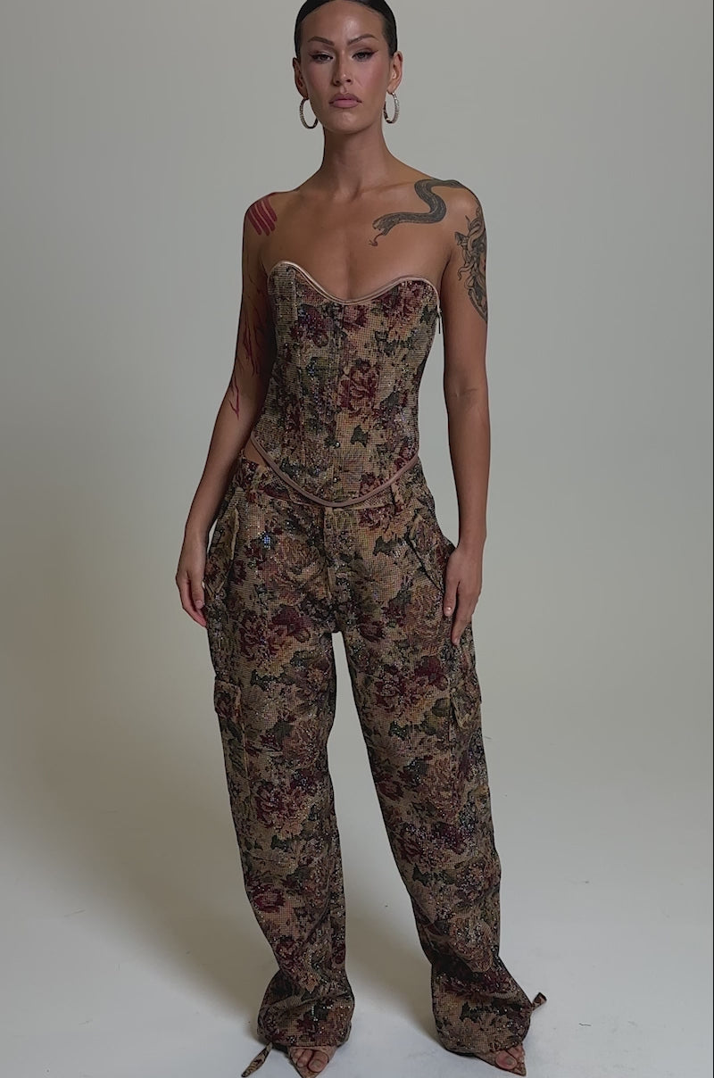 A video of a woman with slicked-back hair, hoop earrings, and visible tattoos poses against a plain background in the GLAM SQUAD RHINESTONE BROCADE CORSET and matching loose floral pants.