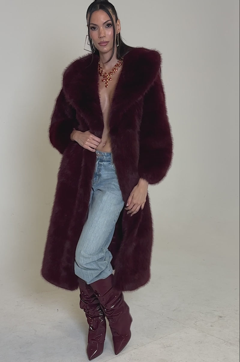 A video of a woman modeling the MANCHESTER HOODED FAUX FUR IN BURGUNDY—a long, deep red jacket with a wide collar—paired with matching knee-high boots and a statement necklace, her hair slicked back against a plain white background.