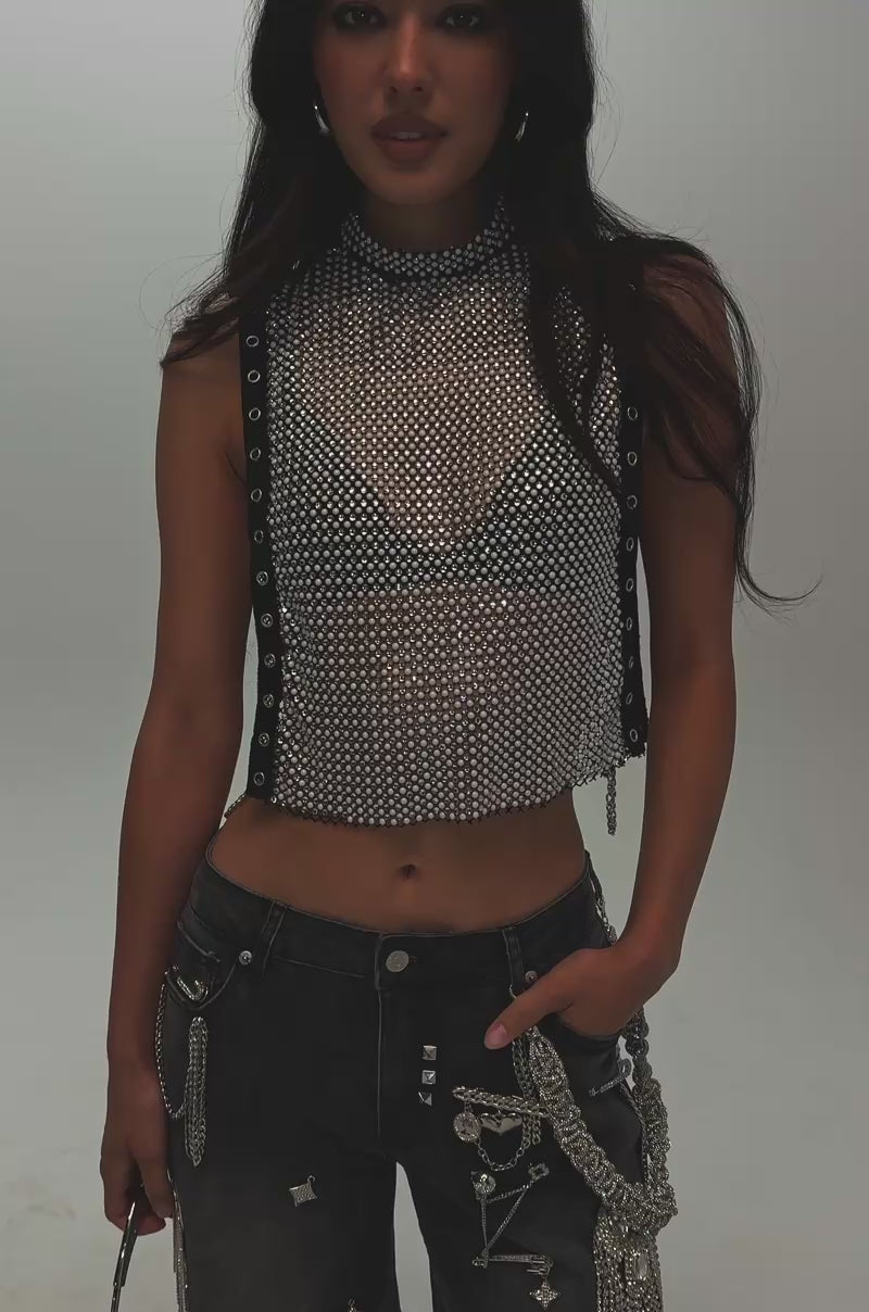 Video of a woman modeling the NO CRYING IN THE CLUB RHINESTONE MESH TOP—a sleeveless crop mesh statement piece layered over a black bra and paired with dark jeans adorned with silver chains and pins, perfect for a striking night-out look.