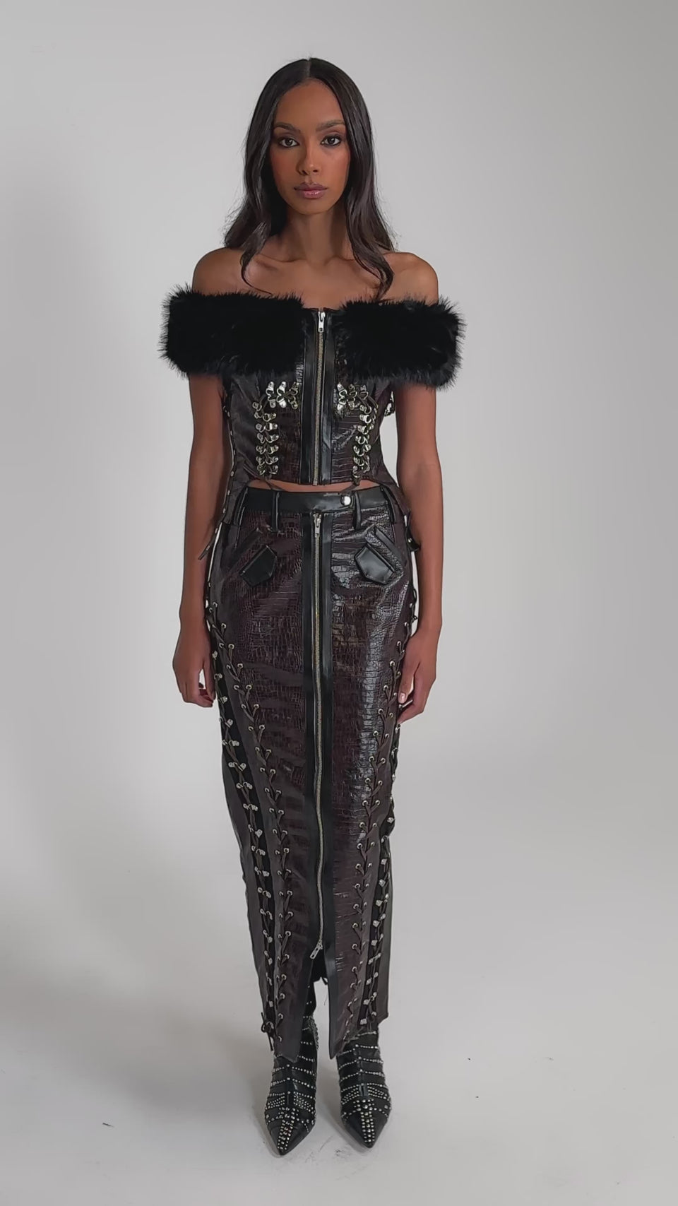 Video of woman modeling the TOO SEXY FOR YOU MAXI SKIRT, a black faux leather maxi with lace-up and zipper details, paired with a black off-the-shoulder, faux fur-trimmed corset top and pointed studded boots.