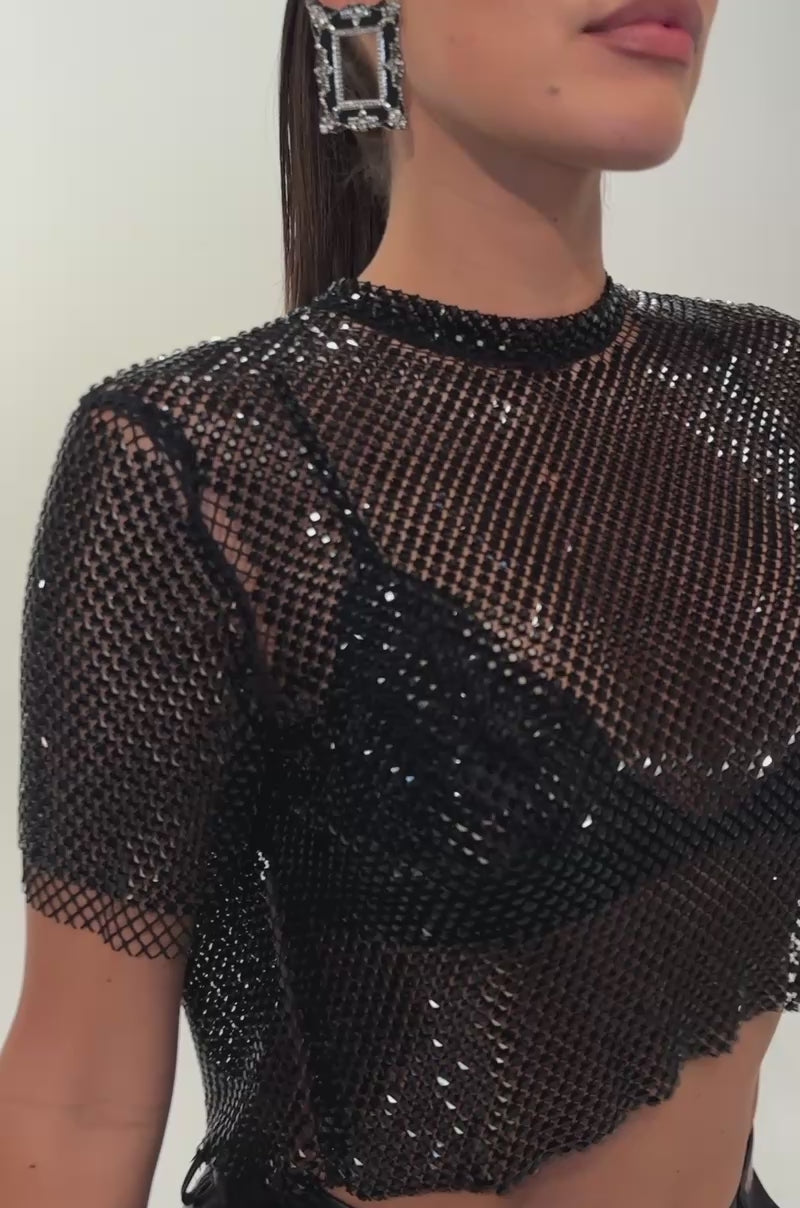 A video of a woman wearing the JUST A FEELING RHINESTONE TEE IN BLACK, styled as a party top layered over a black bra and paired with high-waisted black leather shorts. 