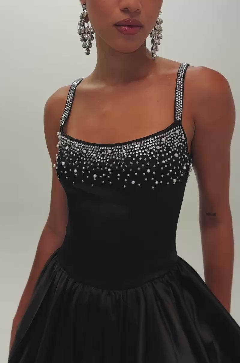 Video of woman modeling the COSMIC DREAM EMBELLISHED MINI DRESS, a sleeveless black mini with rhinestones on the bodice, accessorized with statement earrings and a small handbag.