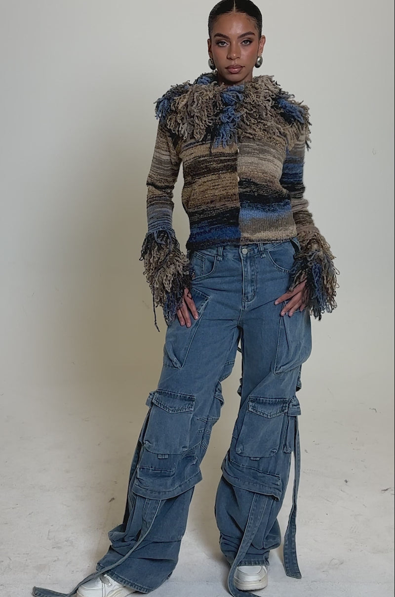 A video of a woman in the ALWAYS ON MY MIND FRINGE SWEATER with frayed edges and ombre design, paired with oversized blue cargo jeans featuring multiple pockets and straps, and white sneakers against a plain light background.
