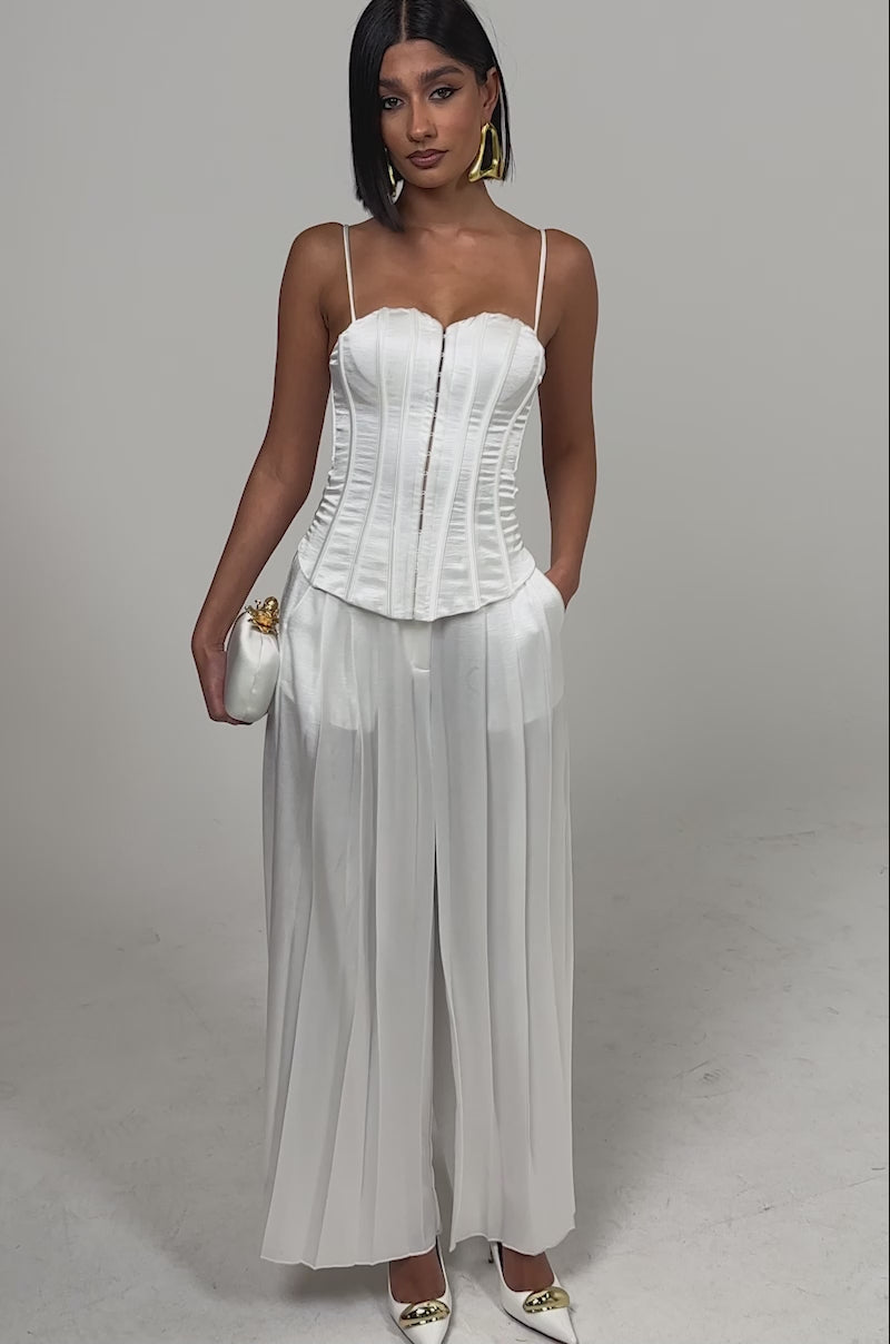A video of a woman wearing the IRIS SATIN CORSET, styled with high-waisted wide-leg white pants, pointed-toe heels, gold earrings, and a small white clutch.