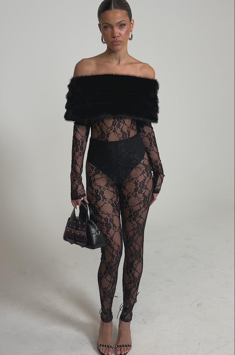 A video of a woman wearing the LACE N FUR SLEEVELESS JUMPSUIT styled with a faux fur shawl, high-waisted briefs, strappy heels, and a black handbag. Her hair is pulled back.