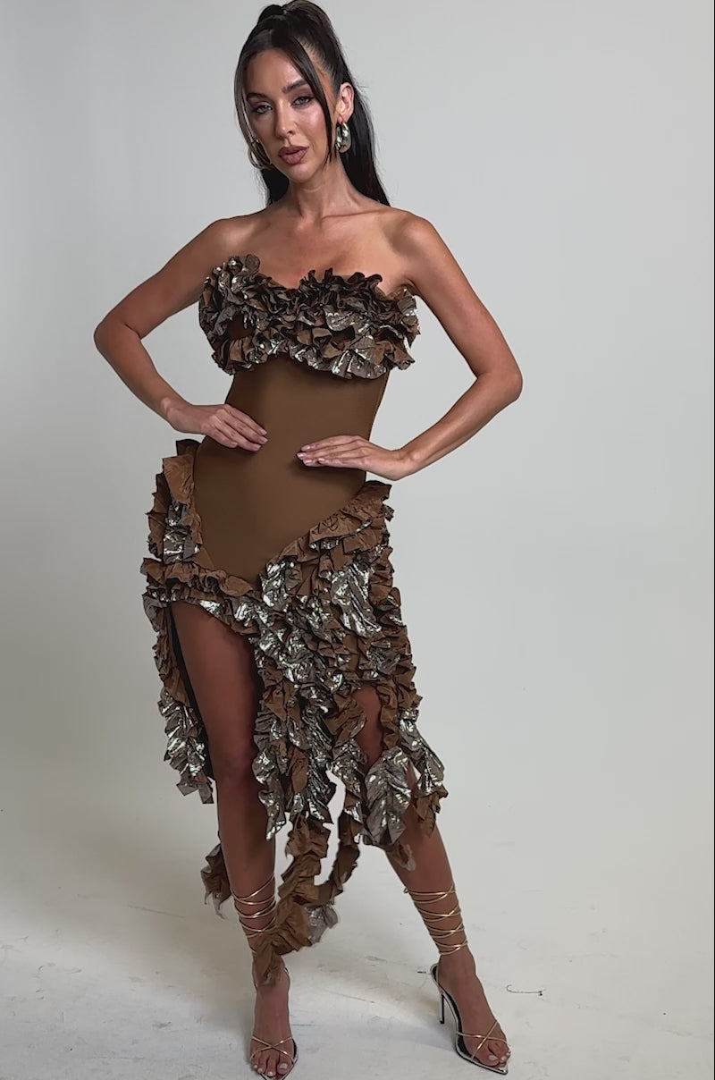 A video of a woman in the CITY LIGHTS RUFFLE MIDI DRESS IN BRONZE, featuring a strapless sweetheart neckline and ruffled asymmetrical hem, styled with strappy heels.