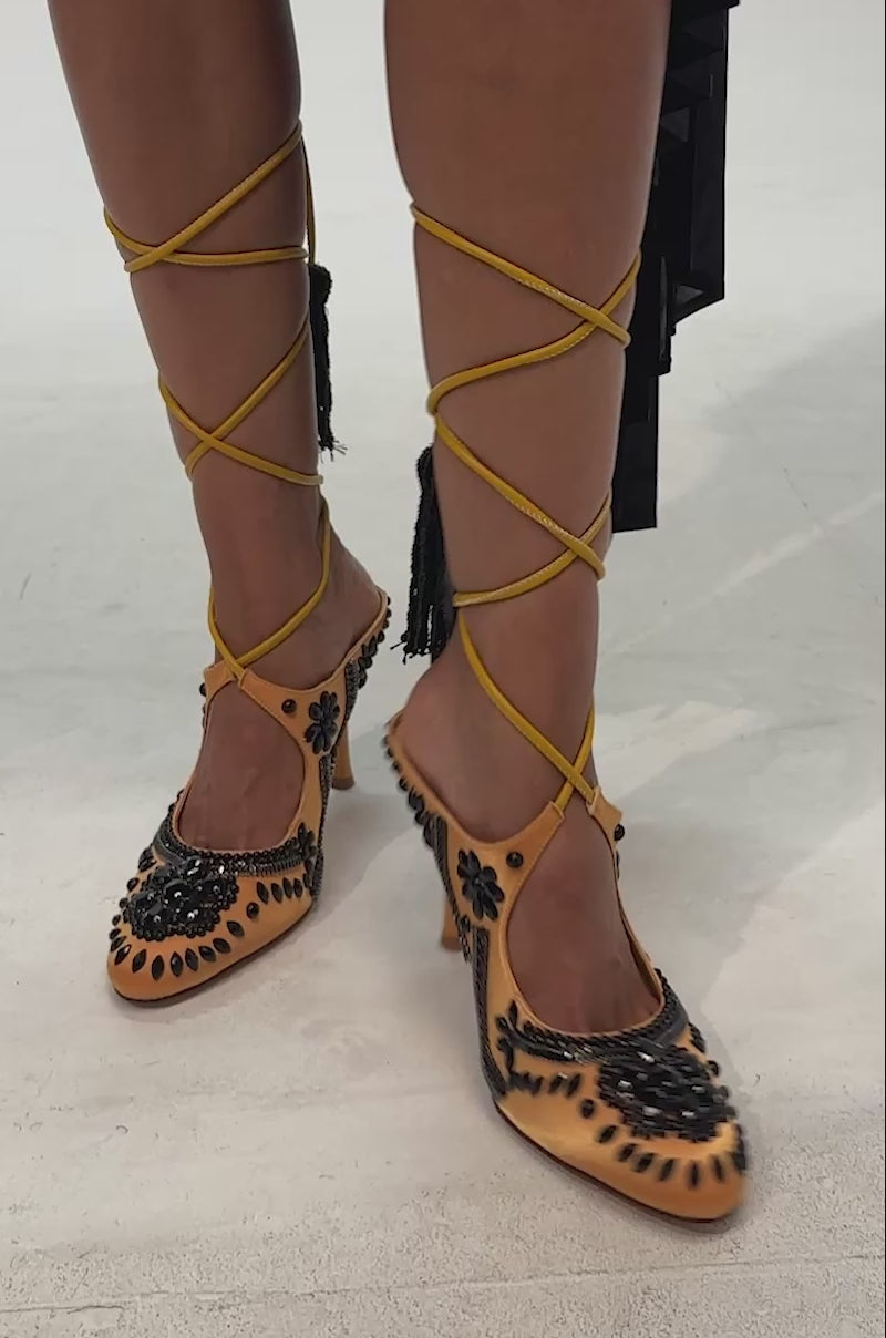 Video of woman modeling the AZALEA WANG ADORN MARIGOLD EMBELLISHED STILETTO PUMP, a yellow faux leather pump adorned with black embroidery, long yellow lace-up straps that wind around the calves
