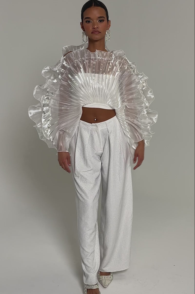Video of woman modeling the SHINING DOWN ON YOU BLOUSE with architectural ruffles and texture, paired with high-waisted white wide-leg pants. Her hair is slicked back, she wears statement earrings, and stands against a plain background.
