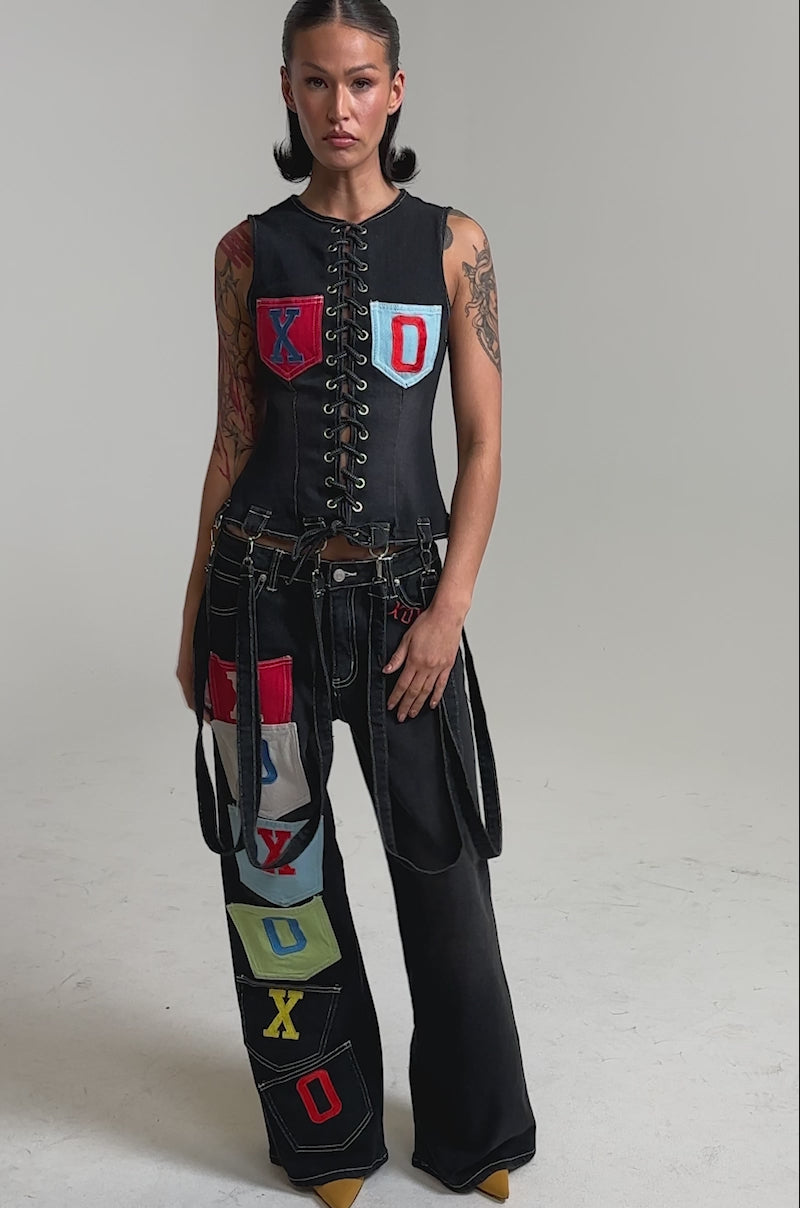 A video of a woman in the BRING IT BACK DENIM PATCHWORK PANT, black wide-leg jeans adorned with colorful "XOXO" letter patches. 